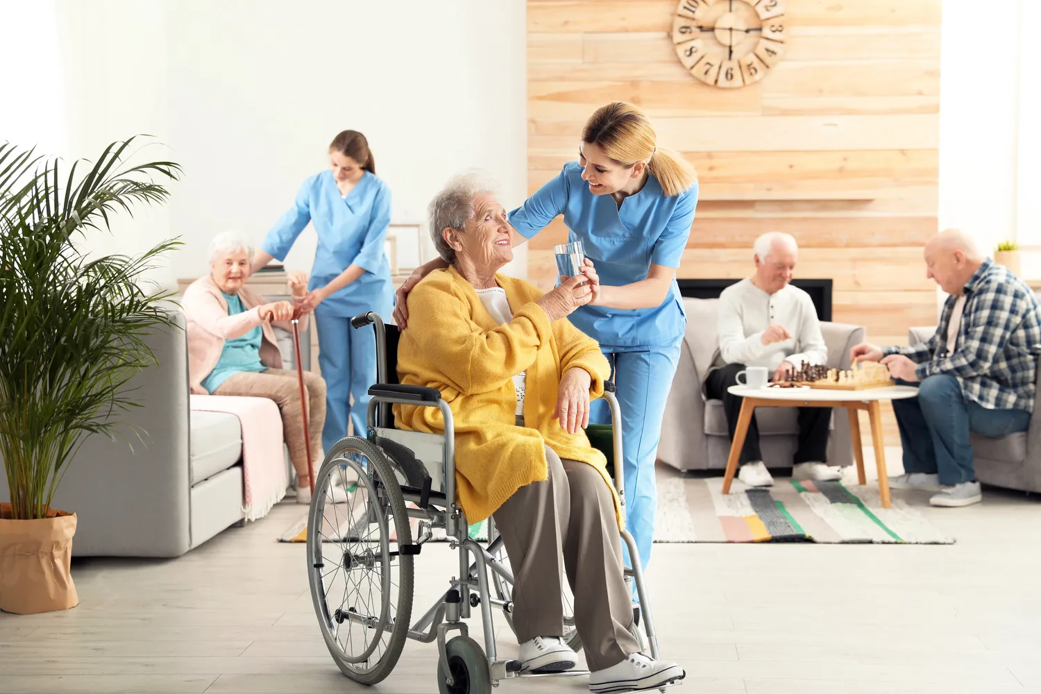 SENIOR GOLD 24 GMBH, Krankenschwester gibt einer älteren Frau im Rollstuhl im Altersheim ein Glas Wasser.